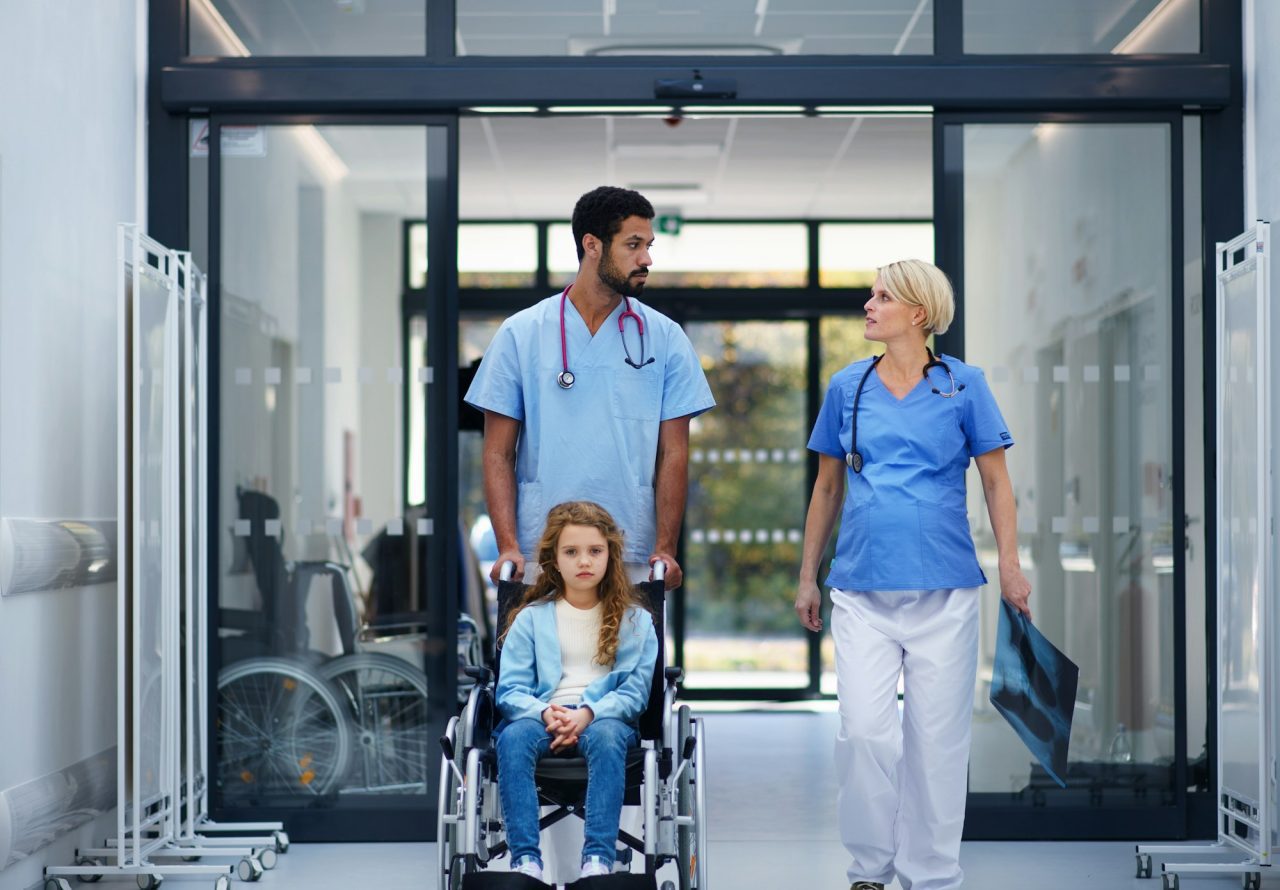 2025 - Início - Home pregnant doctor with her colleague pushing litle girl at wheelchair at pediatric department Experienced and Compassionate Team